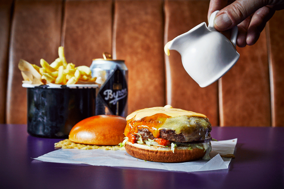 A jug of hot cheese being poured over a stacked juicy burger