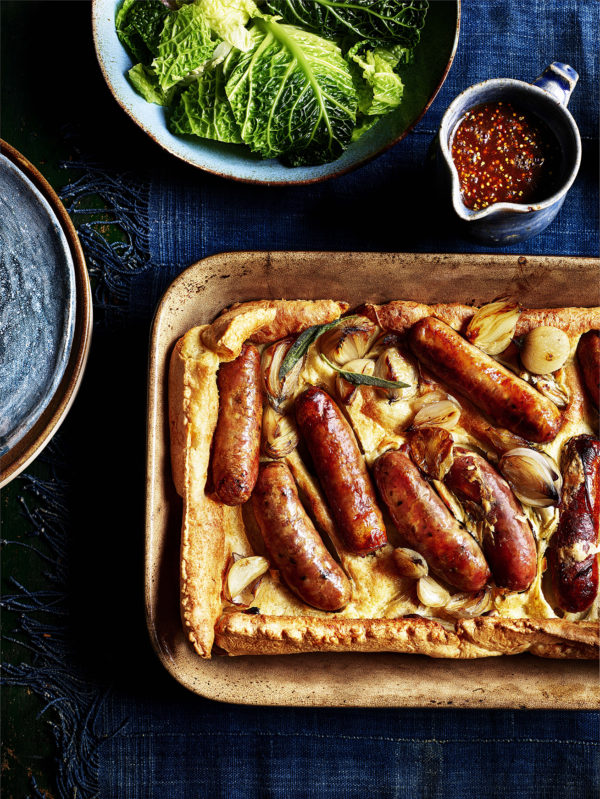Food photography showing a tray of sausages in batter, with cabbage and gravy side dishes