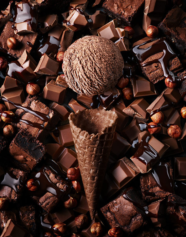 Food and drink photography showing an empty chocolate flavour cone, with a scoop of chocolate ice cream above and surrounded by different types of chocolate