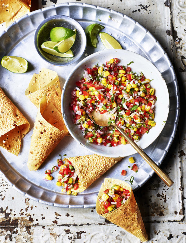 Food product photography showing a bowl of sweetcorn, tomato salsa and several cone shaped indian bread, with a side dish of sliced limes